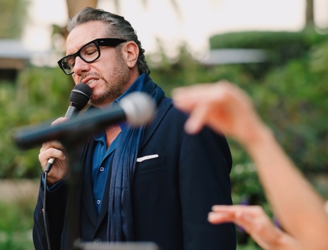 Philippe Harari singing outdoors with microphone and glasses, palm trees in background.