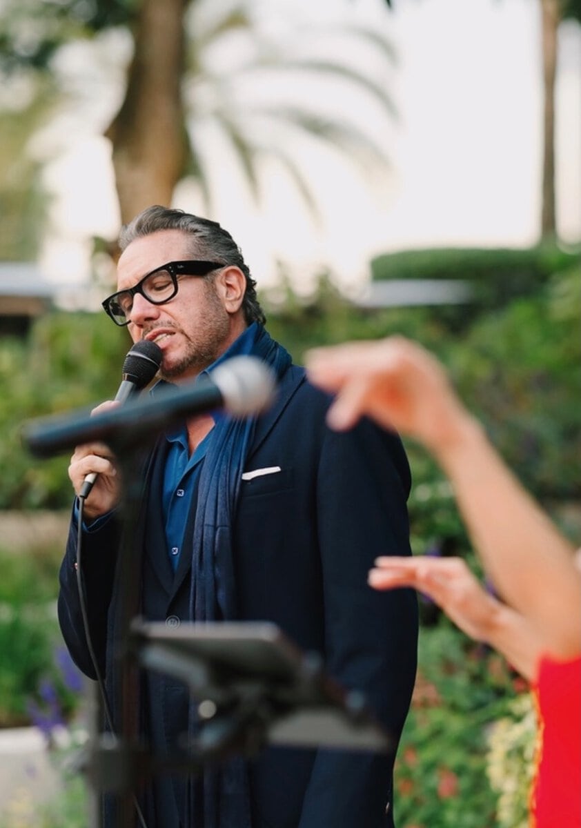 Philippe Harari singing outdoors with microphone and glasses, palm trees in background.